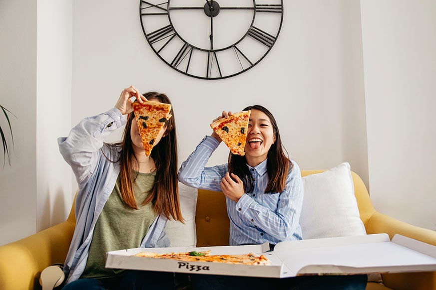 dos chicas sentadas en un sofá con un trozo de pizza en la mano tapándose un ojo.